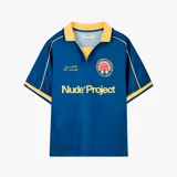 Camiseta de fútbol Nude Project color azul con cuello polo amarillo. Presenta detalles en amarillo en los hombros y mangas, estampado frontal con el nombre de la marca y un escudo circular con una rosa en el pecho.