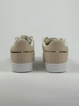 Zapatillas deportivas de cuero sintético en color blanco, beige y negro, con cordones y aplique decorativo dorado con forma de moño.