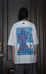 Remera blanca de corte oversize y manga corta, confeccionada en algodón premium. Presenta un estampado gráfico en la espalda con una ilustración estilo cómic de un caballero medieval con una copa y un celular, haciendo referencia a la carta del tarot 'Caballero de copas'.