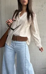 Camisa de manga larga color beige con detalles de encaje y calados verticales en el frente y las mangas, cuello en V y cierre frontal con botones.