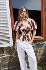 Camisa de rayas verticales rosadas y marrones, con escote en V, mangas corta abullonadas y cierre frontal con múltiples lazadas del mismo tejido. Presenta bajo con acabado de volante.