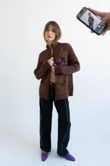 Cardigan de punto color marrón con cierre frontal, cuello alto y bolsillos aplicados. Presenta diseño tejido localizado con detalles en violeta y negro en el pecho.