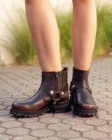 Bota de caña alta confeccionada en cuero color marrón caoba con acabado desgastado. Presenta una suela track robusta, correas con hebillas en la parte superior de la caña y un arnés decorativo con herrajes metálicos en el tobillo.