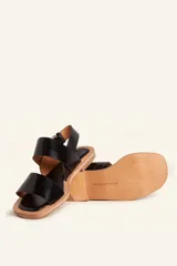 Sandalias planas de cuero negro con tiras anchas en el empeine y cierre ajustable con hebilla metálica en el talón.