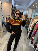 Sweater de punto tejido a mano en mohair, de color negro con diseño de guarda geométrica multicolor en tonos amarillo, naranja y rojo en la zona del canesú. Presenta cuello redondo y mangas largas con puños ajustados.