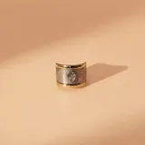 Anillo ancho de diseño moderno, con banda central texturizada y bordes lisos en tono dorado. Presenta una circonia central en forma de gota engastada en una base plateada.