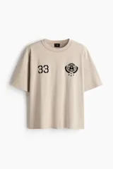 Remera de algodón en punto pesado, color beige, con cuello redondo ribeteado y corte holgado. Presenta estampado frontal con el número 33 y un gráfico circular con estrellas.