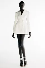 Blazer vestido corto confeccionado en tejido satinado color blanco, con cuello de solapa y cierre frontal cruzado con botones.