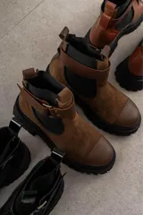 Bota tipo Chelsea de cuero marrón con suela track negra de goma. Presenta paneles elásticos laterales y una costura reforzada en la puntera. Incluye una correa de cuero desmontable con hebilla metálica.