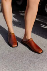 Mules de cuero color suela, con diseño abierto y tira frontal decorada con tachas metálicas circulares. Presentan punta cuadrada y suela plana.