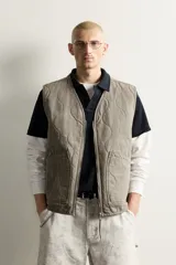 Chaleco de algodón acolchado de corte relaxed fit, color verde militar con efecto lavado. Presenta cuello en V, cierre frontal con cremallera y bolsillos frontales de parche.