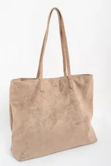 Bolso tipo tote de gran tamaño confeccionado en gamuza sintética color tostado. Presenta un diseño minimalista con asas largas de hombro y una pequeña placa metálica frontal con el logo de la marca.