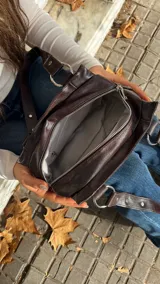 Cartera de hombro color marrón oscuro, confeccionada en material con acabado brillante. Presenta un diseño estructurado con detalles de remaches metálicos plateados en las correas y en el cuerpo de la cartera, además de argollas metálicas que conectan las correas con la base.