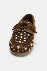 Zapato tipo náutico de serraje color marrón, con detalle de tachas metálicas plateadas en todo el cuerpo y cordones decorativos en la parte delantera. Presenta punta redonda y suela plana.