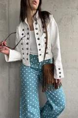 Chaqueta corta de estilo militar en color blanco, con múltiples filas de botones metálicos grabados y detalles de cordones en el frente. Cuenta con cuello mao y mangas largas con botones en los puños.