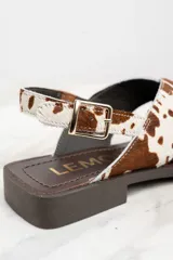 Sandalias bajas con tiras cruzadas de cuero con estampado de vaca en tonos marrón y blanco. Tira trasera con hebilla dorada y suela plana color marrón.
