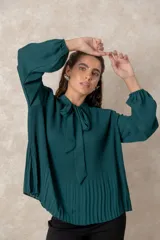 Blusa plisada de color verde oscuro, con mangas largas terminadas en puños elásticos y cuello alto con lazo frontal para anudar.