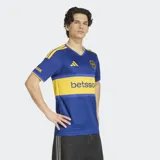 Camiseta de futbol Adidas de la primera equipacion de Boca Juniors, de color azul con la caracteristica franja horizontal amarilla en el pecho. Presenta cuello en V, mangas cortas con las tres tiras de Adidas en los hombros y el logo de la marca y el escudo del club bordados en el frente.
