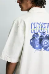 Camisa de corte relajado color verde oliva, con cuello solapa, manga corta y cierre frontal de botones. Presenta un estampado con el texto 'ASTRAL Drift' en el frente.