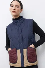 Chaleco capitoneado reversible con cuello alto, cierre frontal con botones a presión y bolsillos delanteros. Presenta un diseño bicolor en tonos verde oliva y beige con detalles en negro.