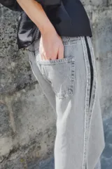 Jean de tiro alto, corte ancho y pierna recta, confeccionado en denim de algodón rígido color gris lavado. Presenta una franja lateral negra contrastante con acabado desflecado en los bordes.