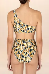 Trikini de una sola pieza con estampado de hojas en tonos mostaza, negro y blanco. Presenta un diseño asimétrico con un solo hombro y recortes laterales unidos por un adorno metálico dorado con forma de hoja.