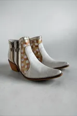 Botas de caña corta de cuero vacuno en tono blanco y detalles metalizados, con diseño pintado a mano en tonos dorados, blancos y terracota en los laterales. Posee taco cuadrado de madera, punta fina y doble cierre lateral con tiradores metálicos personalizados.