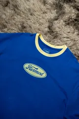 Remera oversize de algodón elastizado, color azul vibrante, con cuello redondo ribeteado en color beige claro. Presenta un estampado frontal ovalado en color beige claro con la palabra "Hermod" en tipografía cursiva.