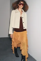Trench corta color beige con cuello solapa, manga larga con trabilla y botón en los puños, bolsillos de vivo en el delantero y cierre frontal con botones oscuros.