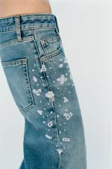 Jeans tiro medio de corte relaxed leg y largo completo, confeccionados en denim azul claro. Presentan detalles de aplicaciones de pedrería y joyas a contraste en los laterales superiores y bolsillos delanteros, estilo clásico de cinco bolsillos y cierre frontal con cremallera y botón metálico.