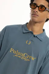 Remera de manga corta color verde oscuro, estilo polo con cuello y abertura frontal. Presenta un bordado central con el texto "PulauCrew" y un laurel en la parte superior.