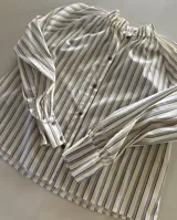 Camisa de rayas verticales, color vainilla con líneas oscuras, cuello alto con volados y botones frontales. Cuenta con mangas largas abullonadas y puños anchos abotonados.