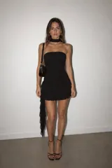Vestido corto negro de estilo strapless con diseño ajustado al cuerpo y falda con volados en capas. Incluye un detalle de cinta tipo choker en el cuello y una caída lateral de tela transparente.