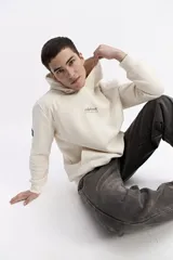 Canguro de felpa color crema, con capucha y bolsillo canguro frontal. Presenta un pequeño estampado con el logo de la marca en el pecho.