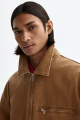 Campera de algodón color marrón con cuello camisero, cierre frontal, bolsillo superior con cierre y bolsillos laterales ribeteados.