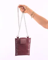 Porta celular de cuero natural color bordó, con diseño rectangular y solapa de cierre. Presenta detalles de tachas metálicas plateadas en los laterales y correa de cadena metálica.