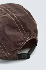 Gorra de visera plana, color marrón oscuro, confeccionada en tejido técnico con costuras contrastantes en blanco. Presenta un parche frontal con un diseño a cuadros y texto.