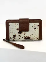 Billetera grande tipo sobre marca Chabas, confeccionada en cuero vacuno con detalle de pelo natural con estampado de manchas tipo vaquita. Cuenta con cierre perimetral, solapa frontal con broche a presión y correa de muñeca extraíble.
