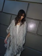 Ruana tejida de diseño abierto con patrón de rayas verticales en tonos crudo y marrón oscuro. Presenta terminaciones con flecos en el ruedo y sutiles detalles de hilo brillante.