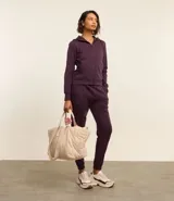 Conjunto deportivo de mujer compuesto por un buzo con capucha y cierre frontal, y un pantalón de jogging a juego. Ambos son de color morado oscuro, con frisos verticales en el centro de las piernas del pantalón.