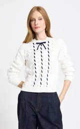 Sweater de punto calado en color blanco, con diseño de trenzas y detalles de cintas azul marino entrelazadas verticalmente en el frente. Presenta cuello redondo, mangas largas con puños acanalados y un pequeño lazo decorativo en el escote.