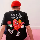 Camiseta negra de algodón con estampado en la espalda de un corazón con zapatillas y flores, con la frase "Love Lifts Us Up" y el logo de Adidas.