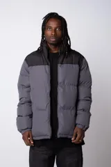 Campera puffer con diseño de bloques de color, combinando negro en la parte superior y gris en el resto de la prenda. Cuenta con cierre frontal completo, cuello alto y puños elásticos.