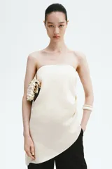 Top strapless de satén color beige, con diseño ceñido, drapeado lateral en la cintura y bajo asimétrico.