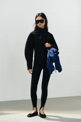 Chaqueta negra de cuello subido con cierre frontal de cremallera termosellada, manga larga con puños anchos y cremalleras laterales en los puños.