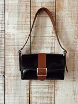 Cartera de cuero negro con brillo charol y detalles en cuero marrón texturado, incluyendo la tira vertical central con hebilla metálica y la correa de hombro ajustable.