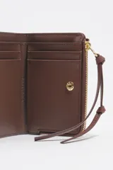 Billetera monedero de piel color marrón de Bimba y Lola, con estructura rígida y estampado de pequeñas palmeras doradas. Cuenta con cierre de cremallera superior con tirador de tela, logo impreso en el frente y tarjetero interior.