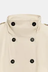 Chaleco de cuello alto color beige con doble botonadura frontal en contraste negro, sin mangas, con trabillas en los hombros y pliegues decorativos en el delantero.