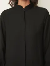 Camisa de manga larga en color blanco, con cuello mao y detalle de volados sutiles en el borde. Presenta cierre frontal oculto con botones y puños con volados.