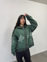 Buzo con capucha y cierre frontal completo, color verde oscuro con efecto lavado degradado. Presenta estampado gráfico en tipografía estilizada en el frente y mangas, bolsillos laterales y etiqueta de marca en el puño.